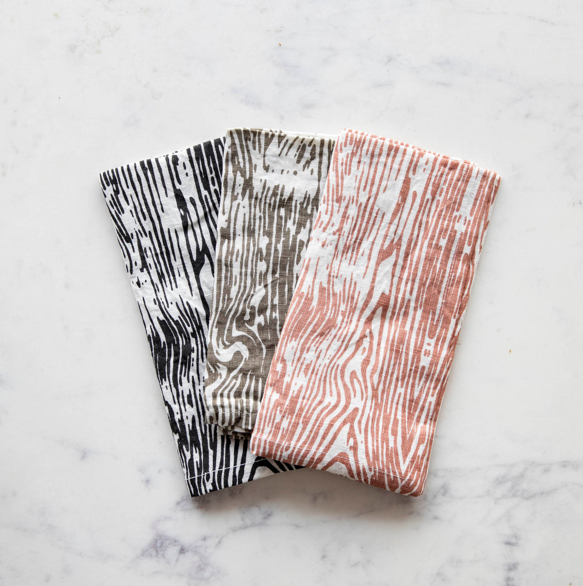 Two linen napkins with a woodgrain pattern, one in black and white color , another in a beige and white color, and the other in a pink hue, displayed on a marble surface.