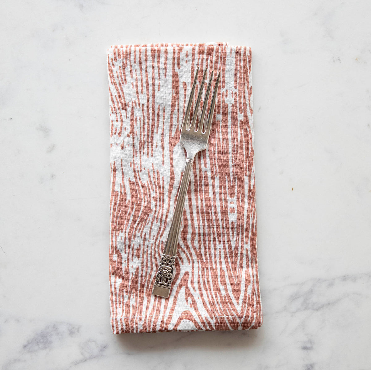 A folded woodgrain-patterned linen napkin in a coral/pink and white color on a white marble surface with a vintage silver-plate fork atop it.