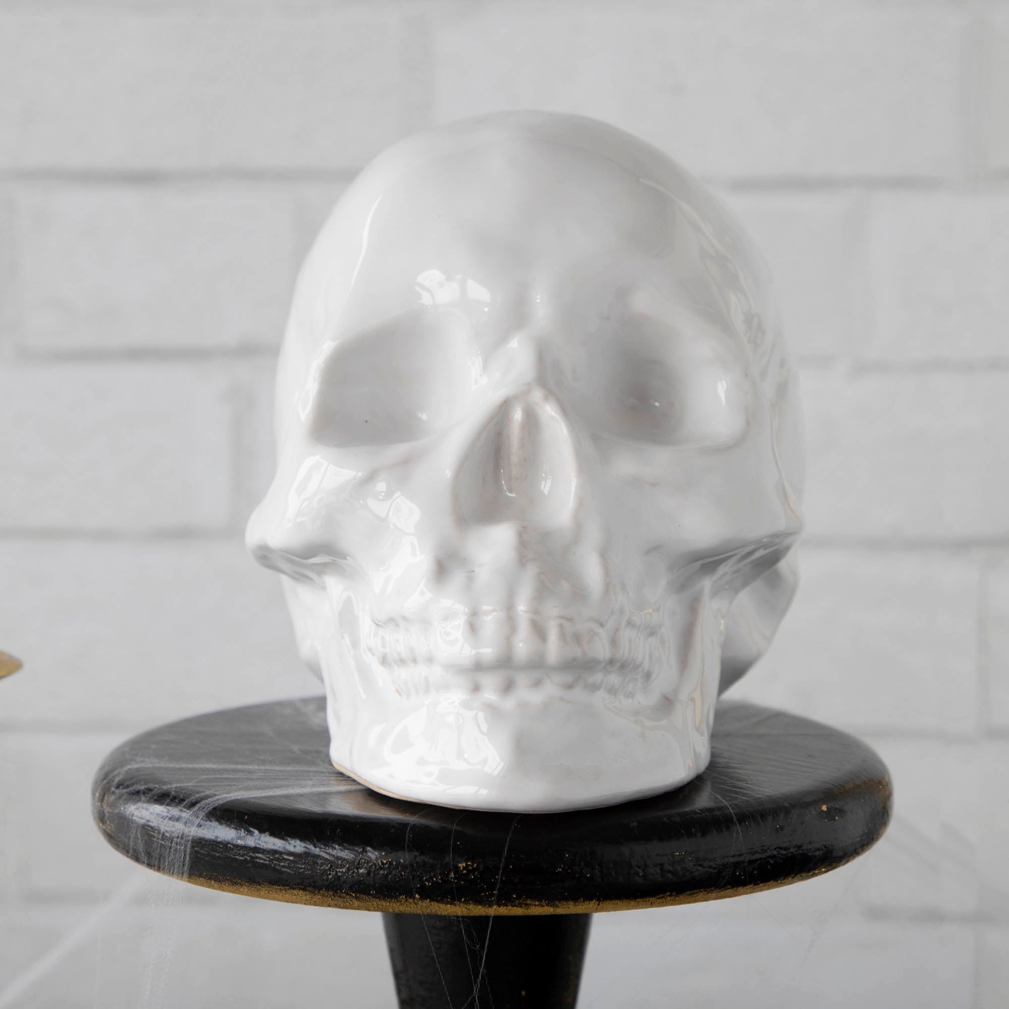Two HomArt ceramic Skull Heads on a striped tablecloth flanked by lit candles in brass holders against a white brick background, serving as an ominous decor piece.