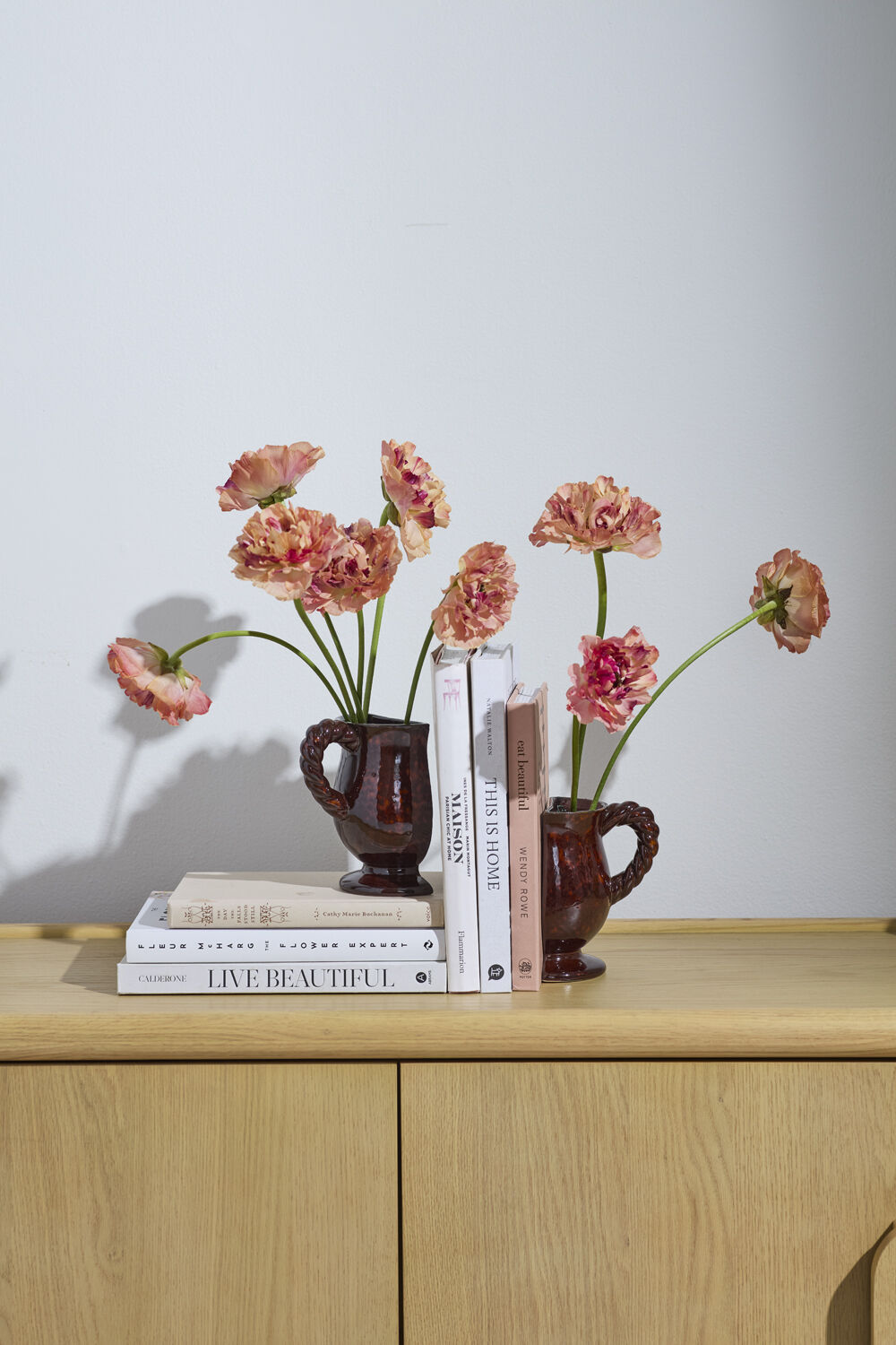 Two small brown vases with pink flowers on a wooden surface against a light gray wall.