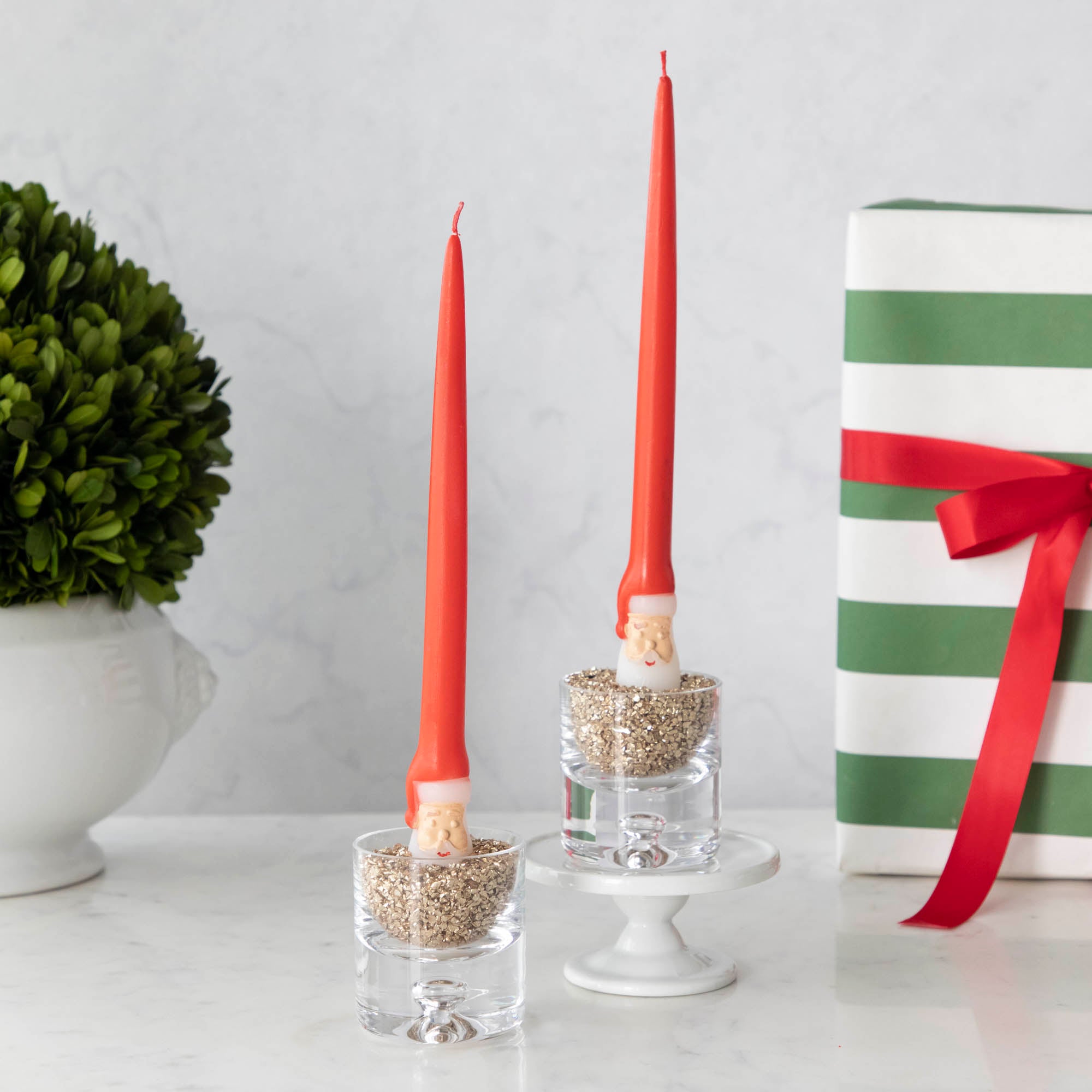 Two taper candles with Santa Claus designs on them, placed in clear candle holders, with one holder sitting on a white stand. The candles are red with white accents and Santa Claus face details.