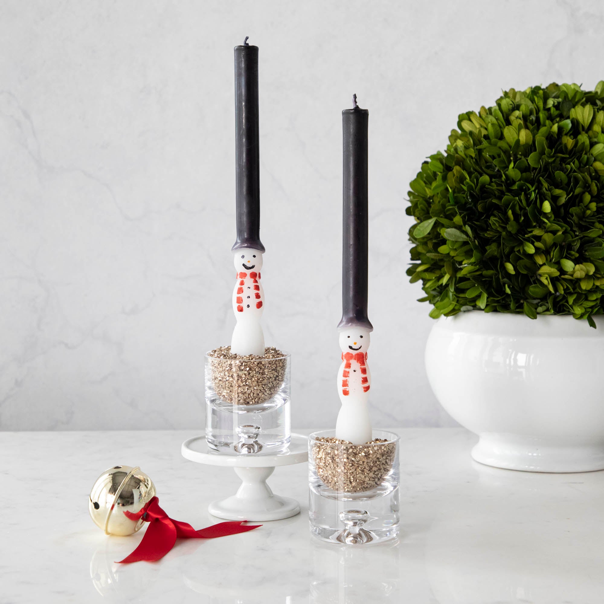 Two taper candles designed to look like snowmen with black hats, red scarves, and white bodys, set on a white surface with a white plant pot in the background and a red bell decorative item to the left.