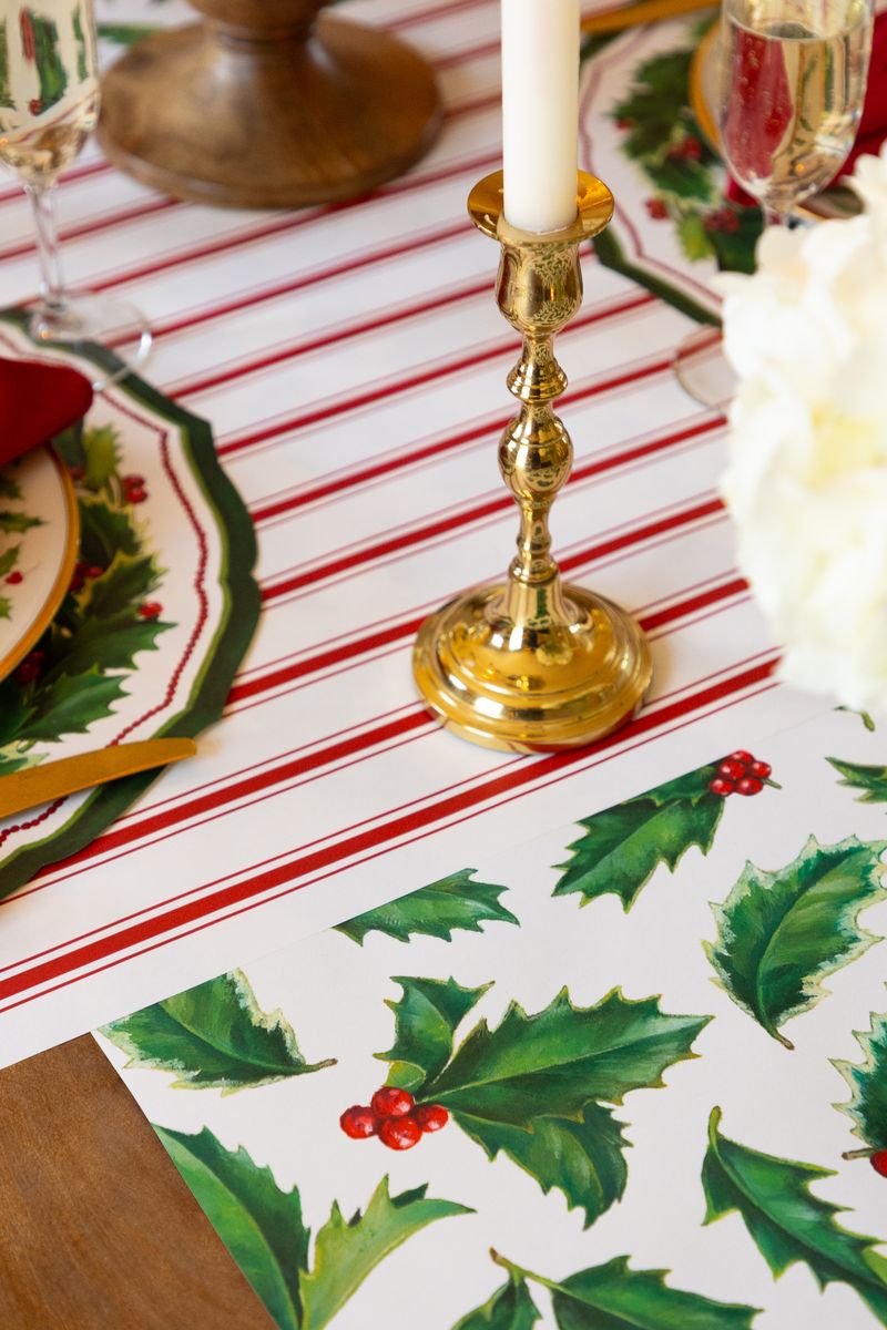 Christmas-themed table setting with a holly designed table runner, gold candlestick, and red and white striped table runner.