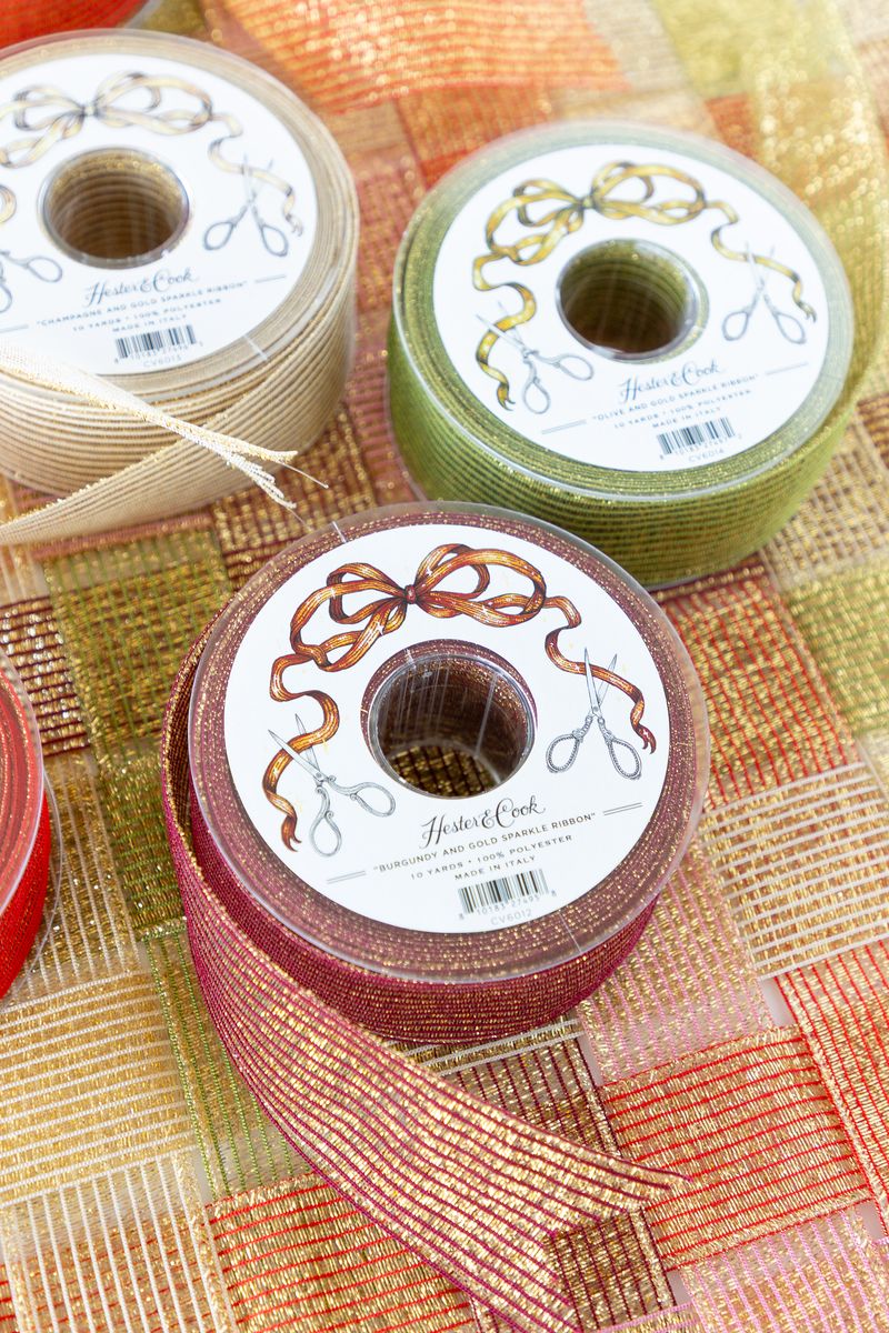 A spool of sparkly burgundy and gold ribbon on a surface of woven sparkle ribbon in five different colors.