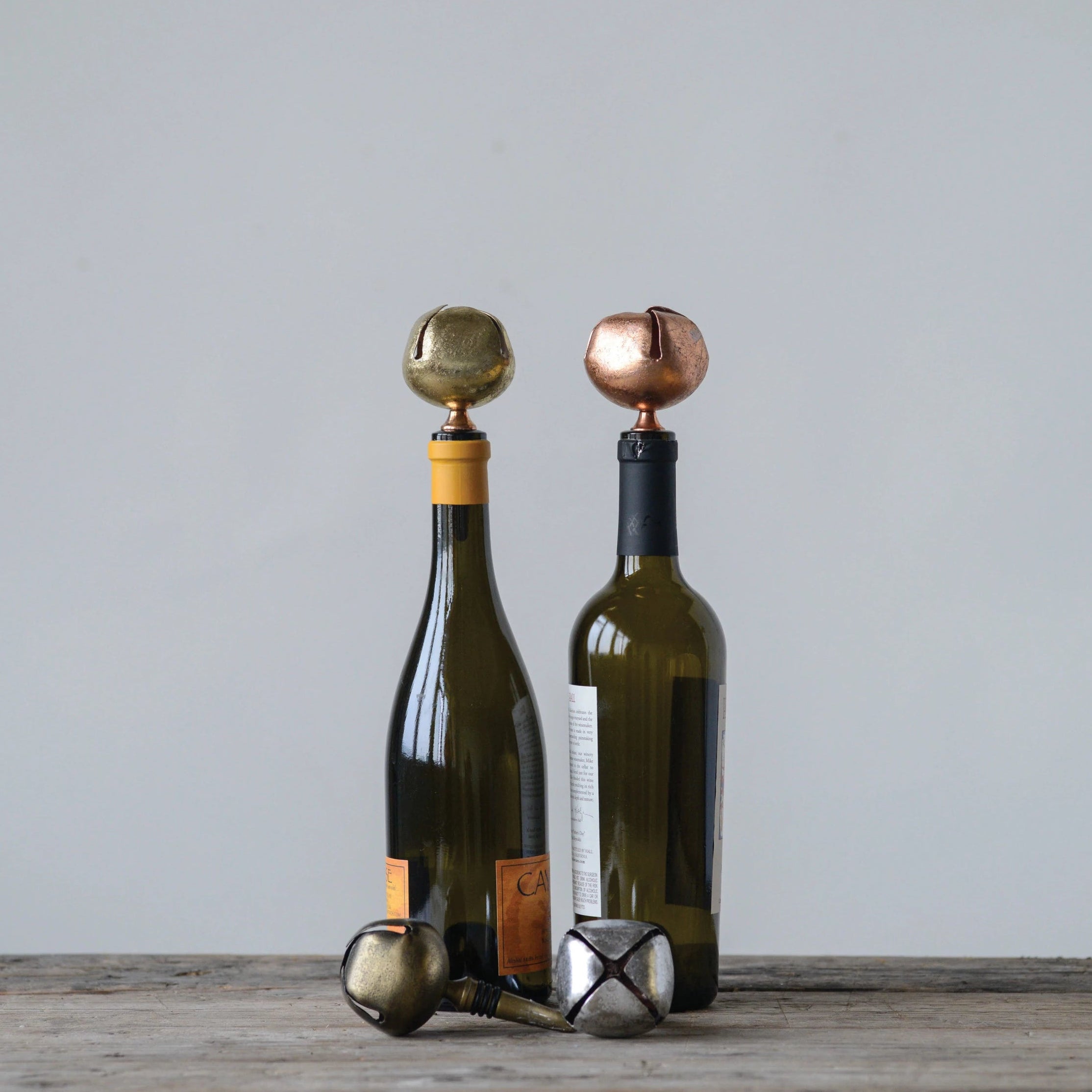 Four metal bottle stoppers with bell designs, in silver, bronze, gold, and black colors, two in bottles.