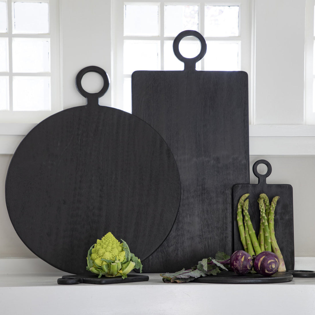 A set of three Be Home Blackened Serving Boards of different shapes and sizes with fresh vegetables displayed in front of a window.