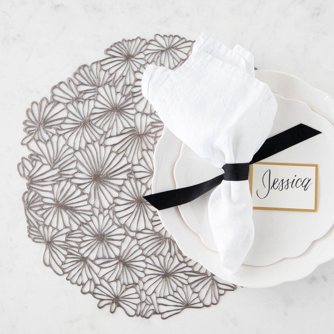 Elegant table setting with a decorative Chilewich Pressed Daisy Mat charger placemat, white porcelain dishes, a folded napkin, and a name card for Jessica.