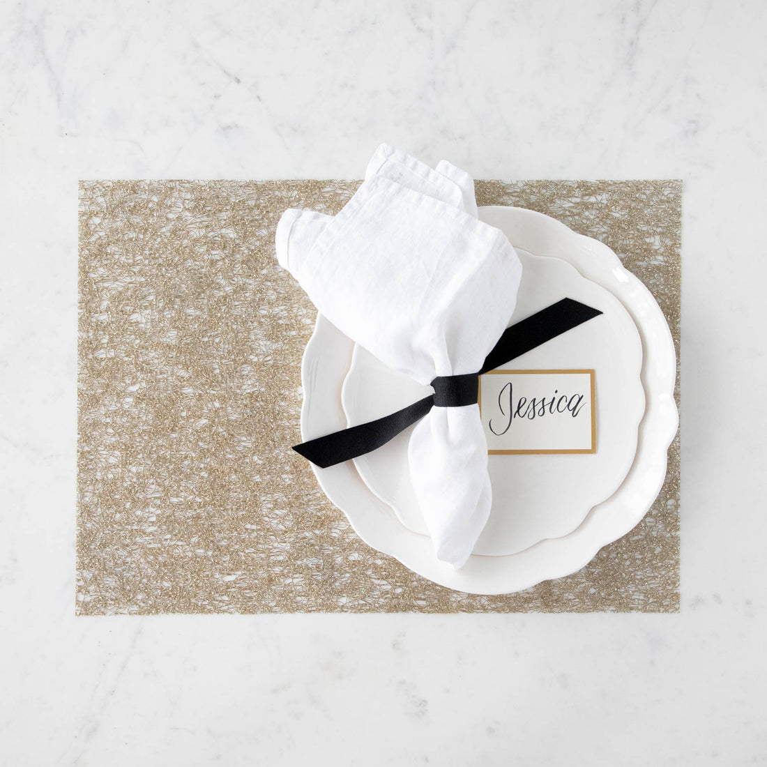 White napkin on a plate with a name card reading &quot;Jessica&quot; at a table setting on a glittery gold Chilewich Metallic Lace placemat.