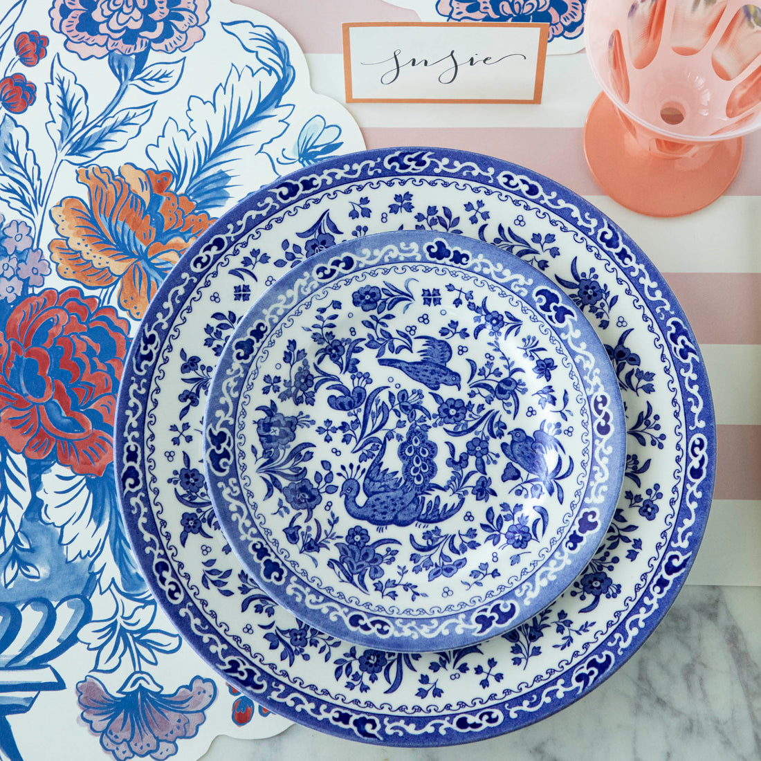 Blue and white patterned Burleigh Blue Regal Peacock Dinnerware plates on a floral patterned background with a name card and a pink glass vase.
