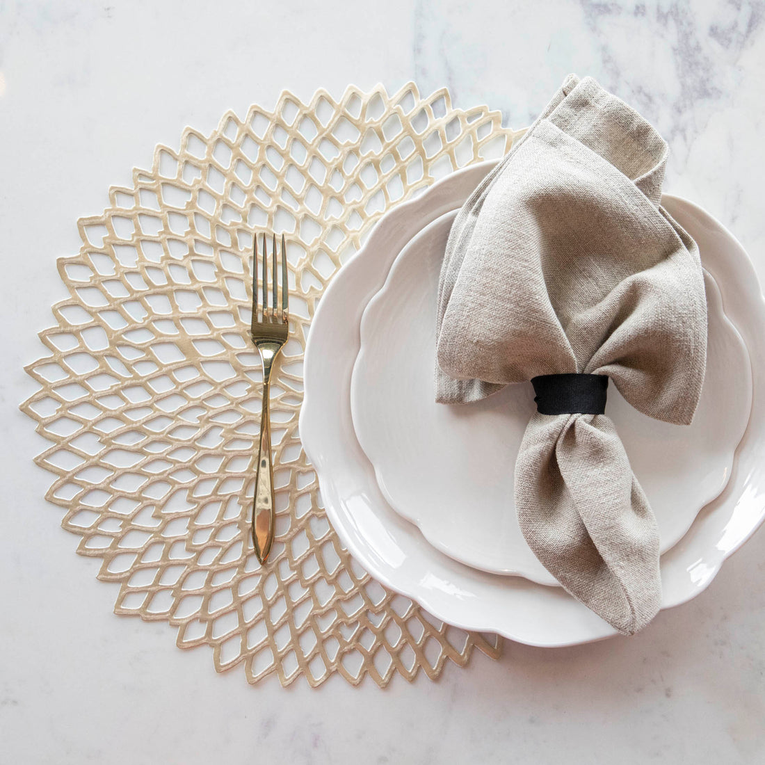 An elegantly set table with a decorative gold Chilewich Pressed Dahlia Mat, white dinner plate, a folded napkin with a ring, and gold silverware.