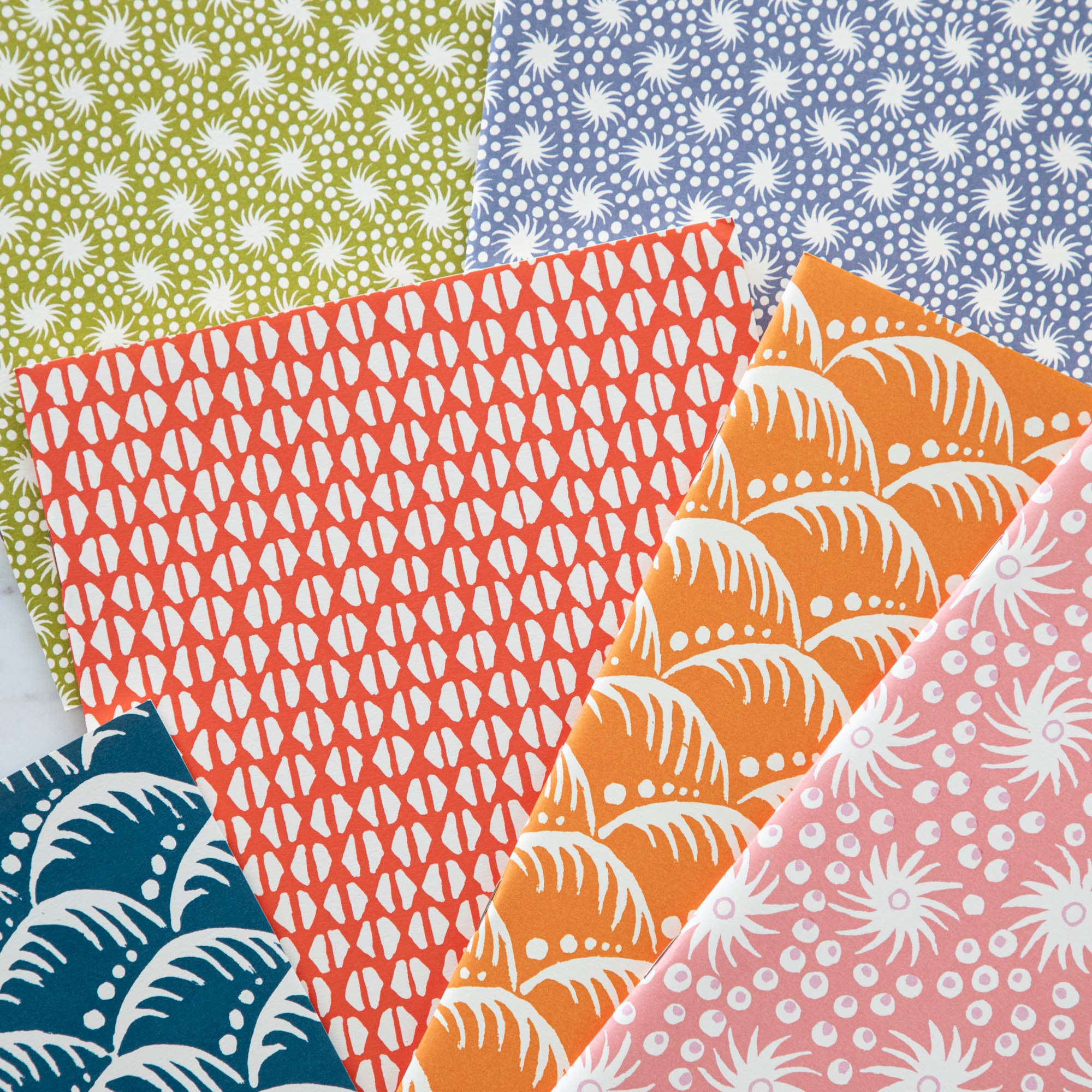 Five colorful Cambridge Imprint Blank Notebooks with patterned covers and three pencils on a marble surface.