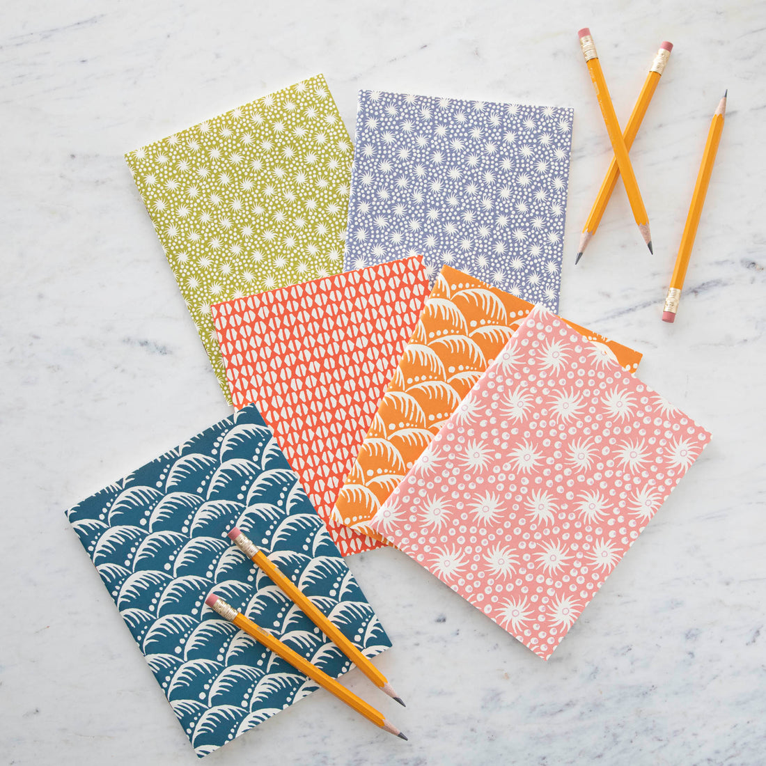 Five colorful Cambridge Imprint Blank Notebooks with patterned covers and three pencils on a marble surface.