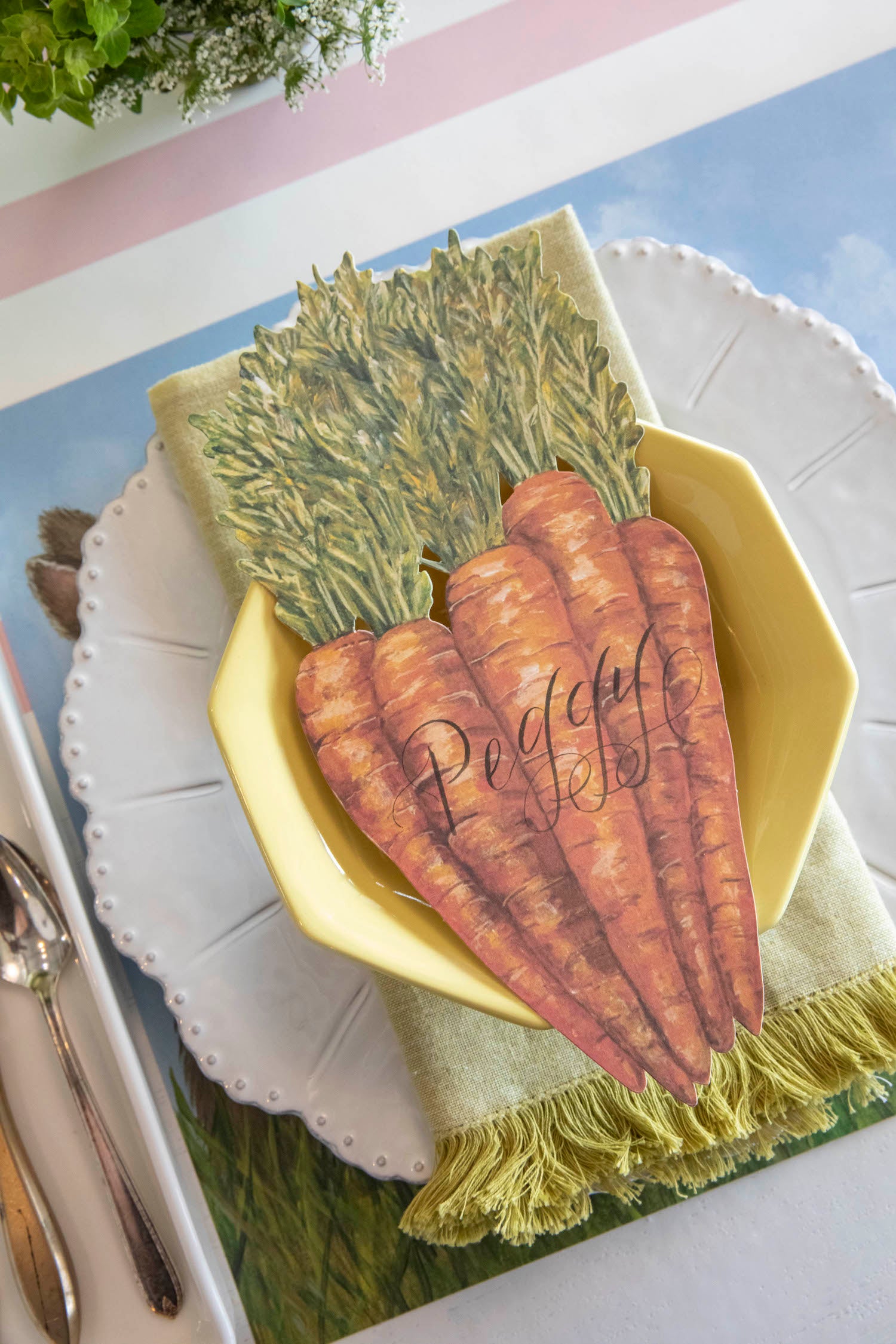 A Carrots Table Accent labeled &quot;Peggy&quot; resting on the plate of a springtime place setting, from above.