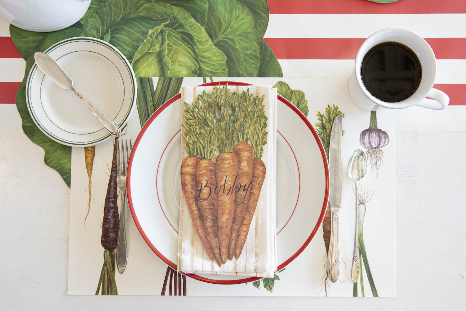 A Carrots Table Accent labeled "Bobby" resting on the plate of a vegetable-themed place setting, from above.