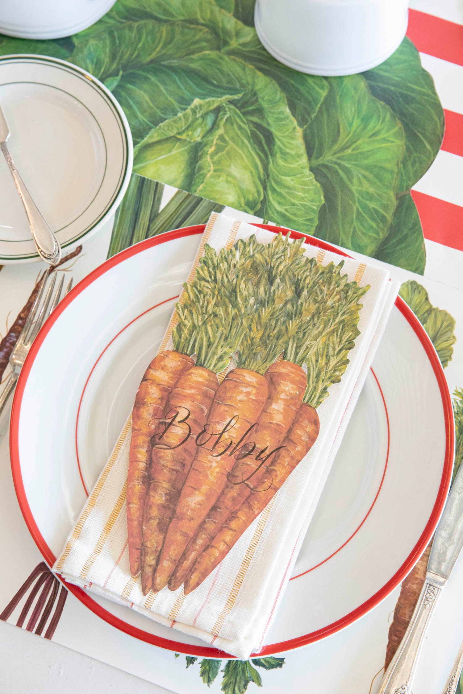 A Carrots Table Accent labeled "Bobby" resting on the plate of a vegetable-themed place setting, from above.