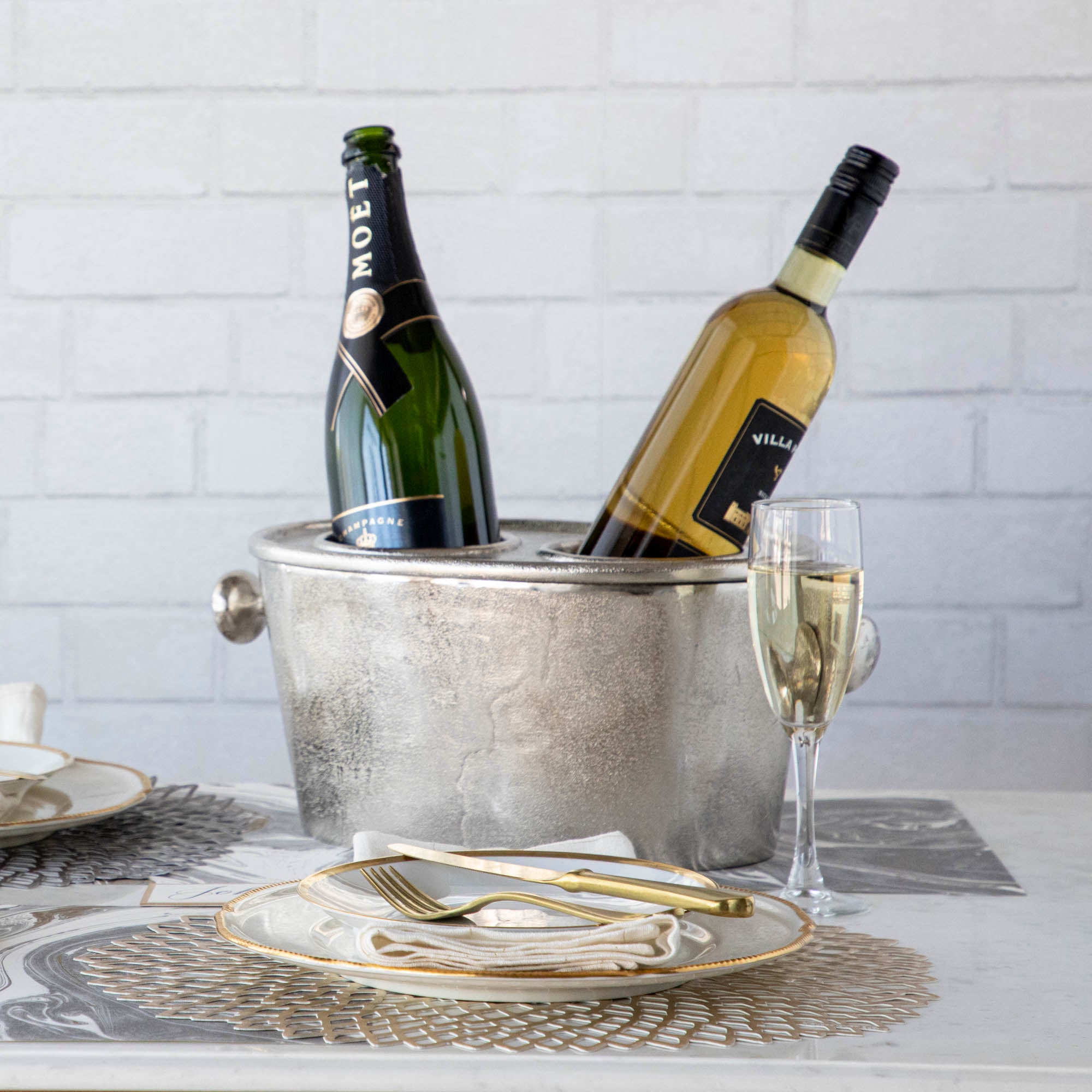 A silver aluminum wine holder with two bottles of wine placed inside, on a table set with a plate, glass, and cutlery.