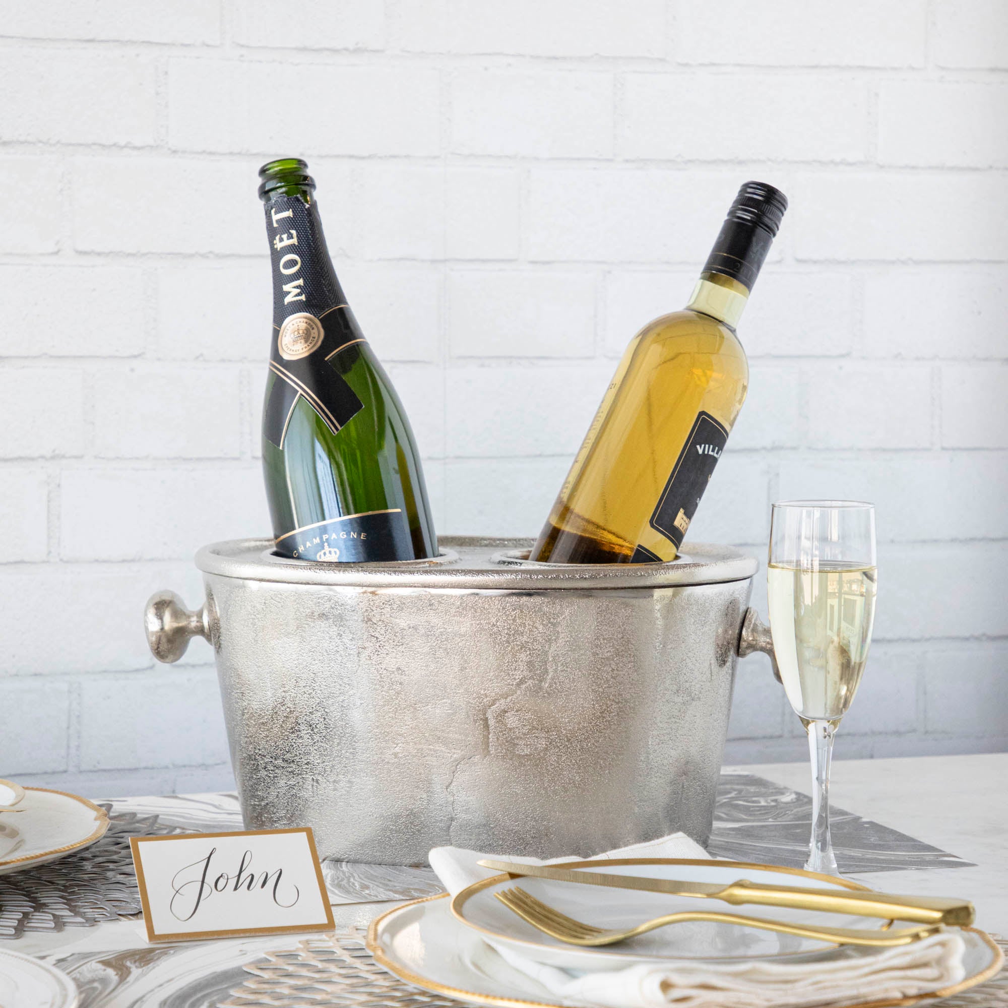 A silver aluminum wine holder with two bottles of wine placed inside, on a table set with a plate, glass, and cutlery.