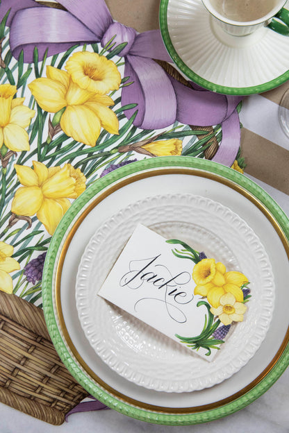 An illustrated wicker basket with a big purple ribbon bow on the handle, full of vibrant yellow daffodils and purple blooms with green foliage, on a white background.