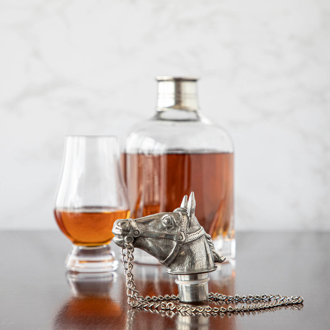 An ornamental crystal Menagerie whisky/spirit decanter, glass, and a Menagerie horse-head stopper on a wooden surface with a marble backdrop.