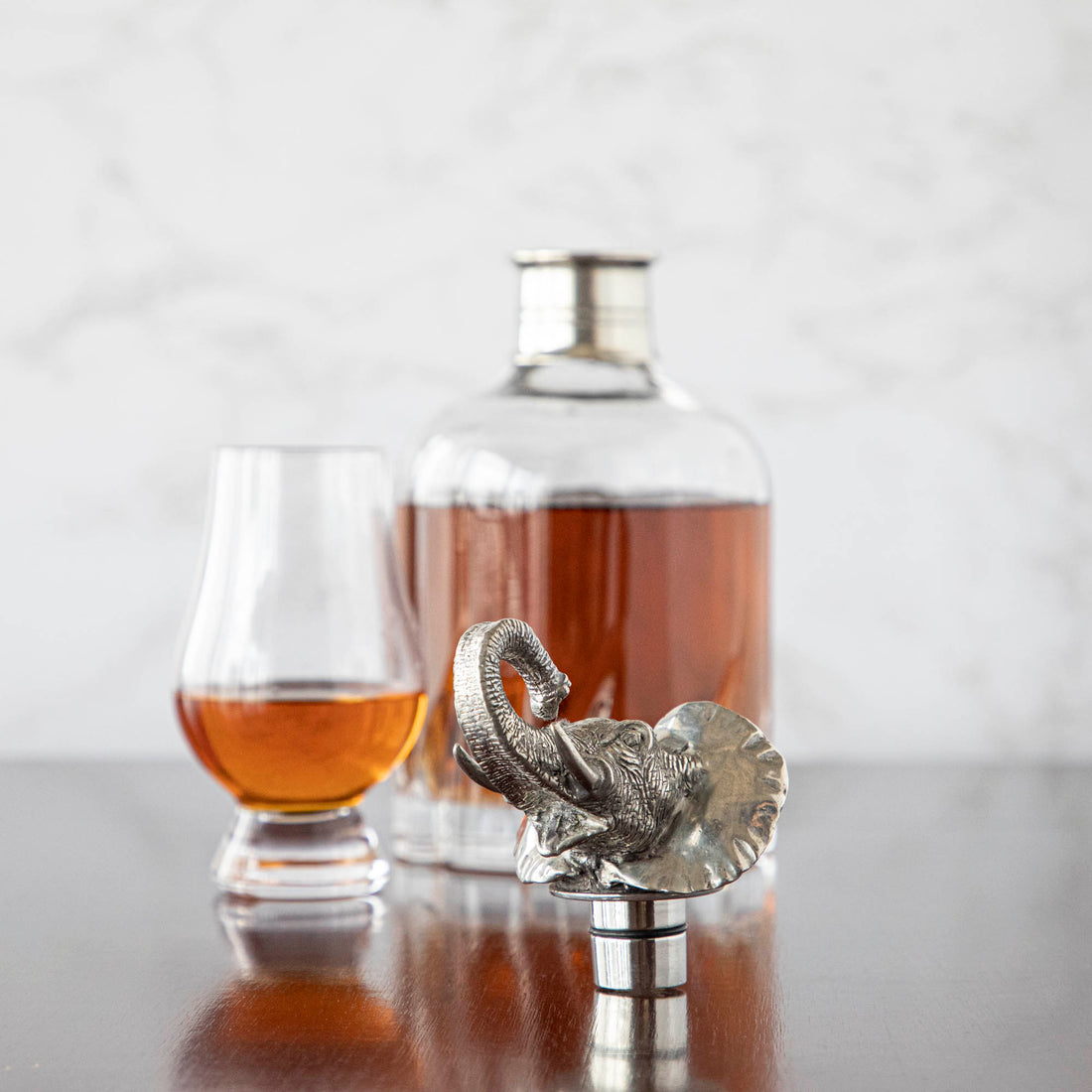 An Menagerie elephant-shaped bottle stopper on a table in front of a glass of whisky and a Menagerie crystal decanter.