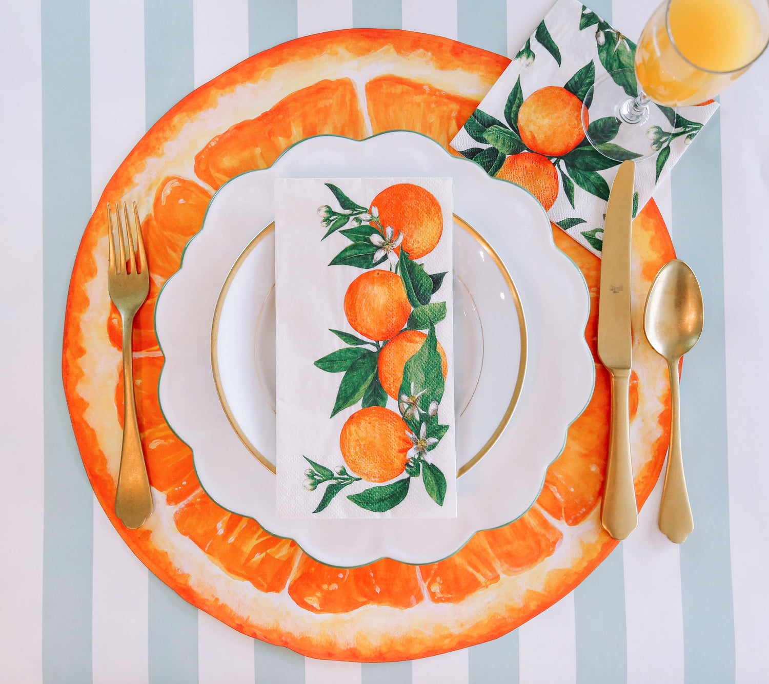 A vibrant citrus-themed place setting, featuring an Orange Orchard Guest Napkin centered on the plate, and an Orange Orchard Cocktail Napkin under a glass of orange juice, from above.