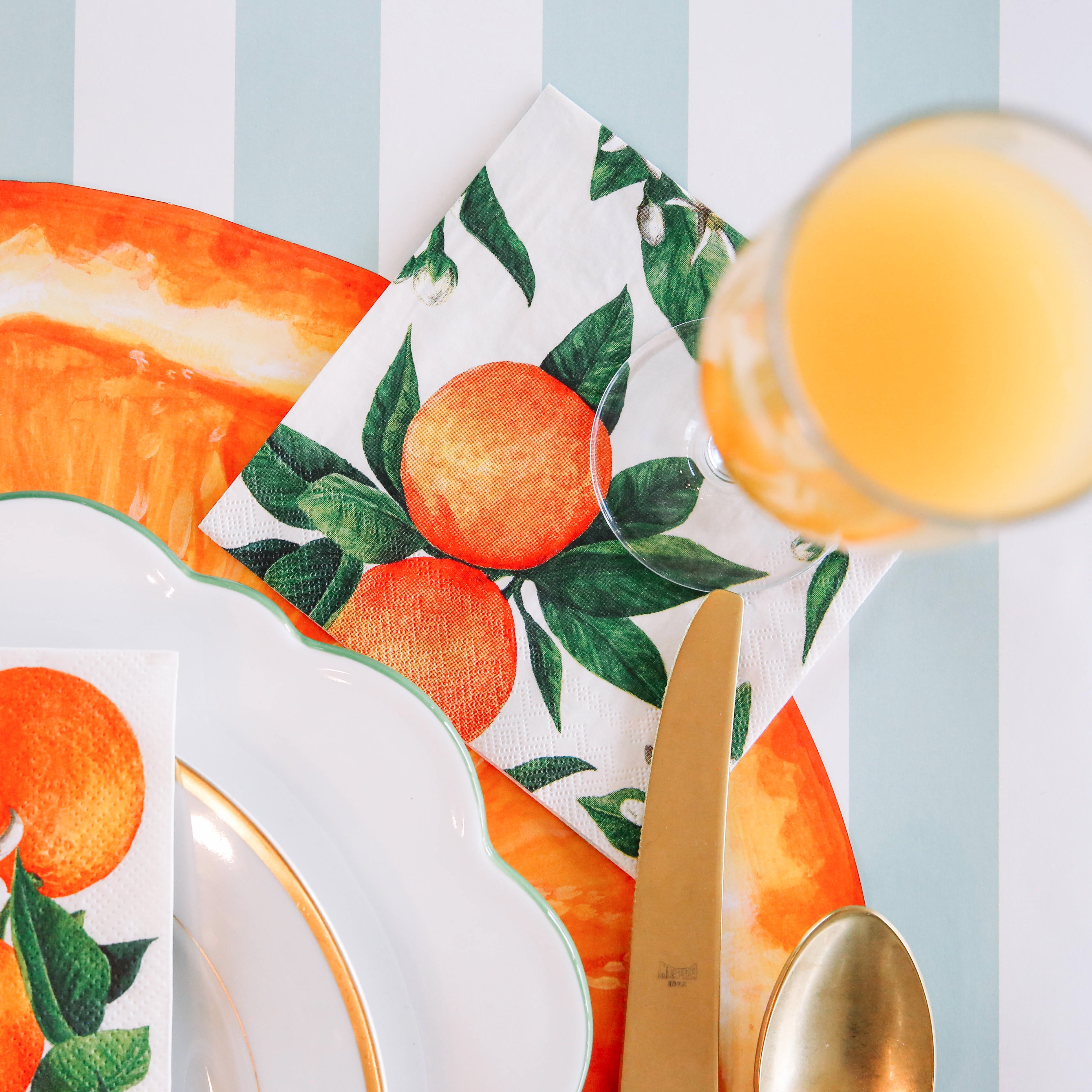 An Orange Orchard Cocktail Napkin under a glass of orange juice in a citrus-themed place setting.