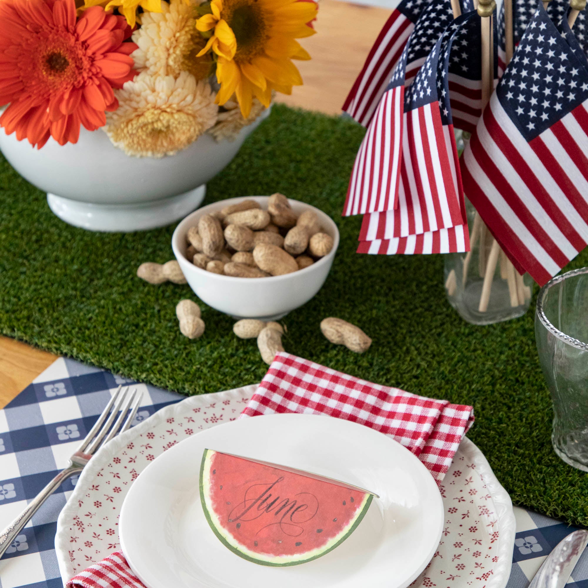 Celebrate the 4th of July with a festive table setting featuring watermelon, sunflowers, and flags. This party setup also includes the addition of Talking Tables Artificial Grass Table Runner as a unique touch.
