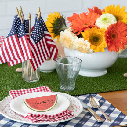 This festive 4th of July party table setting features American flags and a vibrant watermelon centerpiece. The table is adorned with a Talking Tables Artificial Grass Table Runner for added flair.