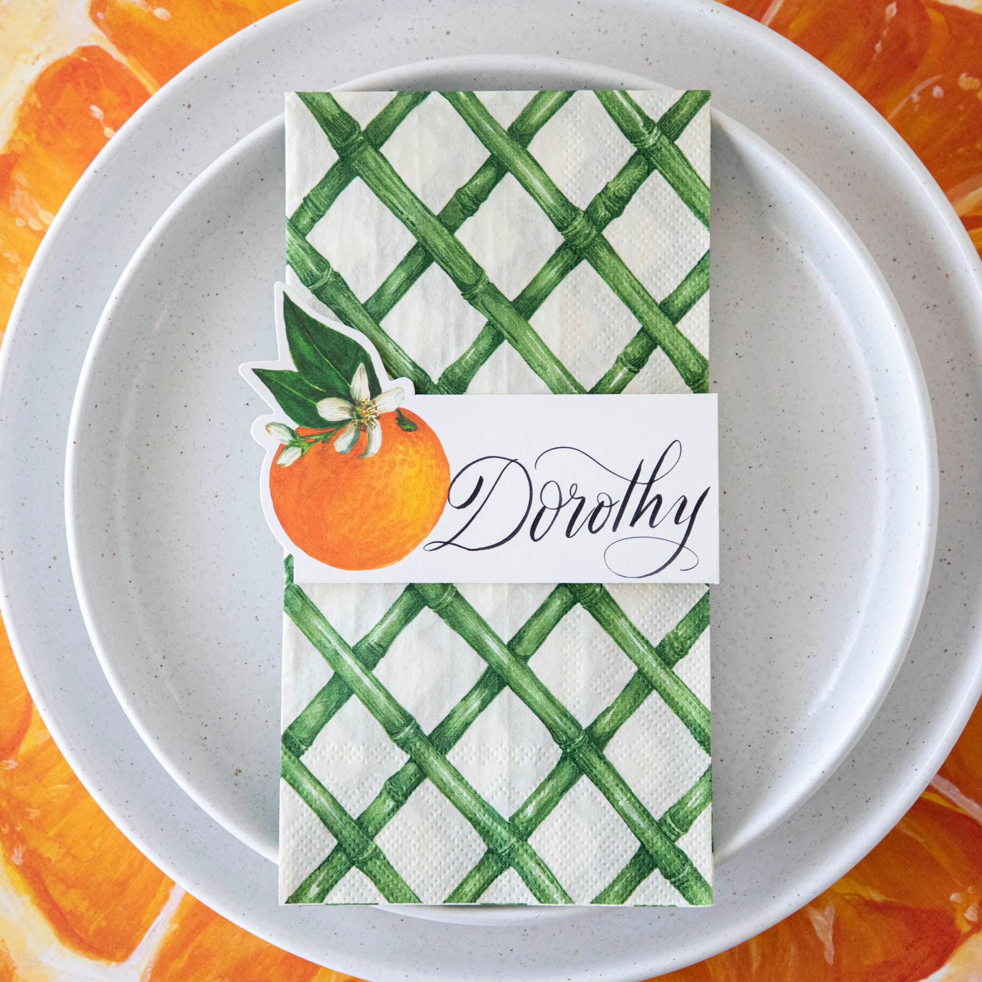 Top-down view of an elegant orange-themed place setting, featuring a Green Lattice Guest Napkin centered on the plate, with a place card.