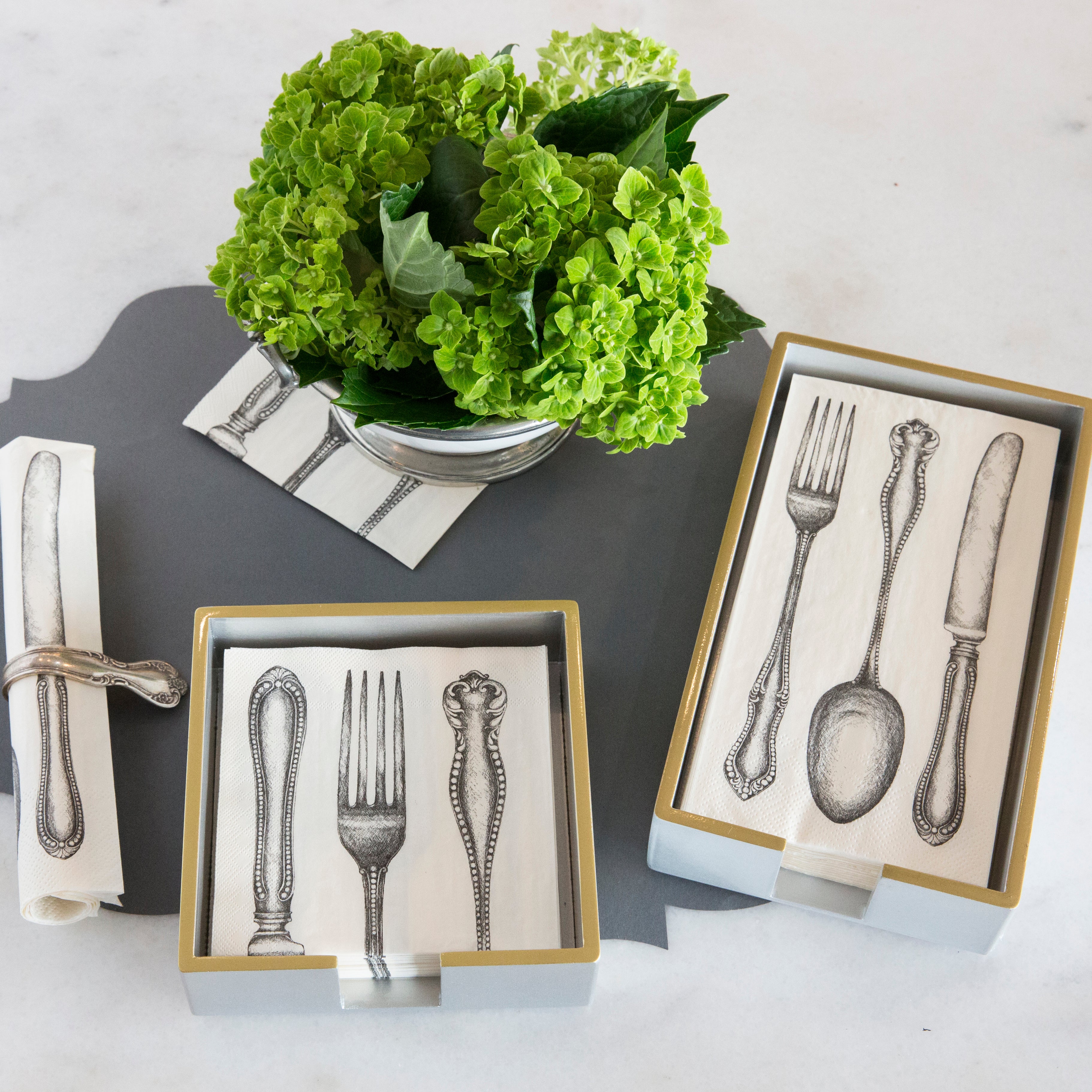 A silver lacquered napkin holder in cocktail and guest size with napkins in them on a grey background and a vase of flowers.