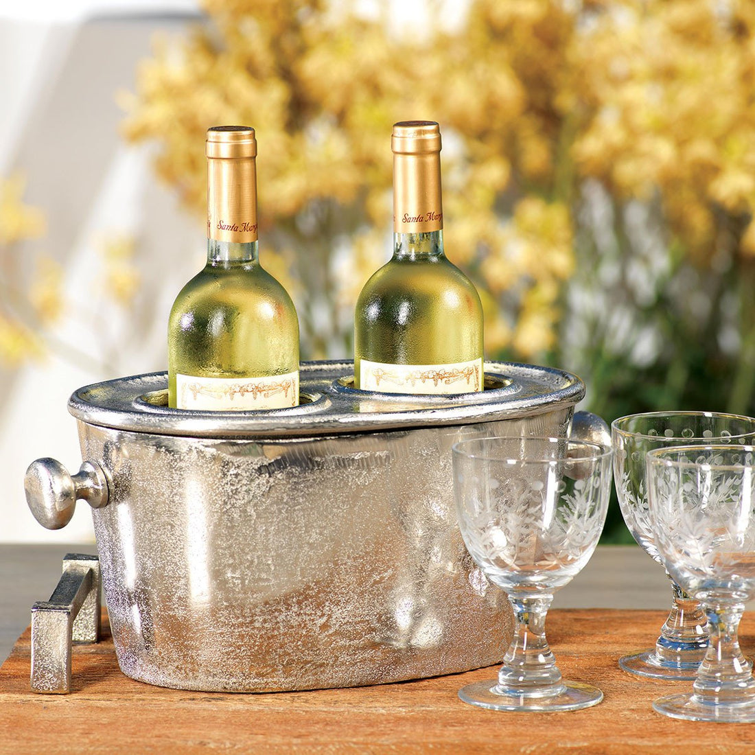 Two bottles of white wine in a Zodax Two Bottle Aluminum Wine Holder with stemware on a wooden table.