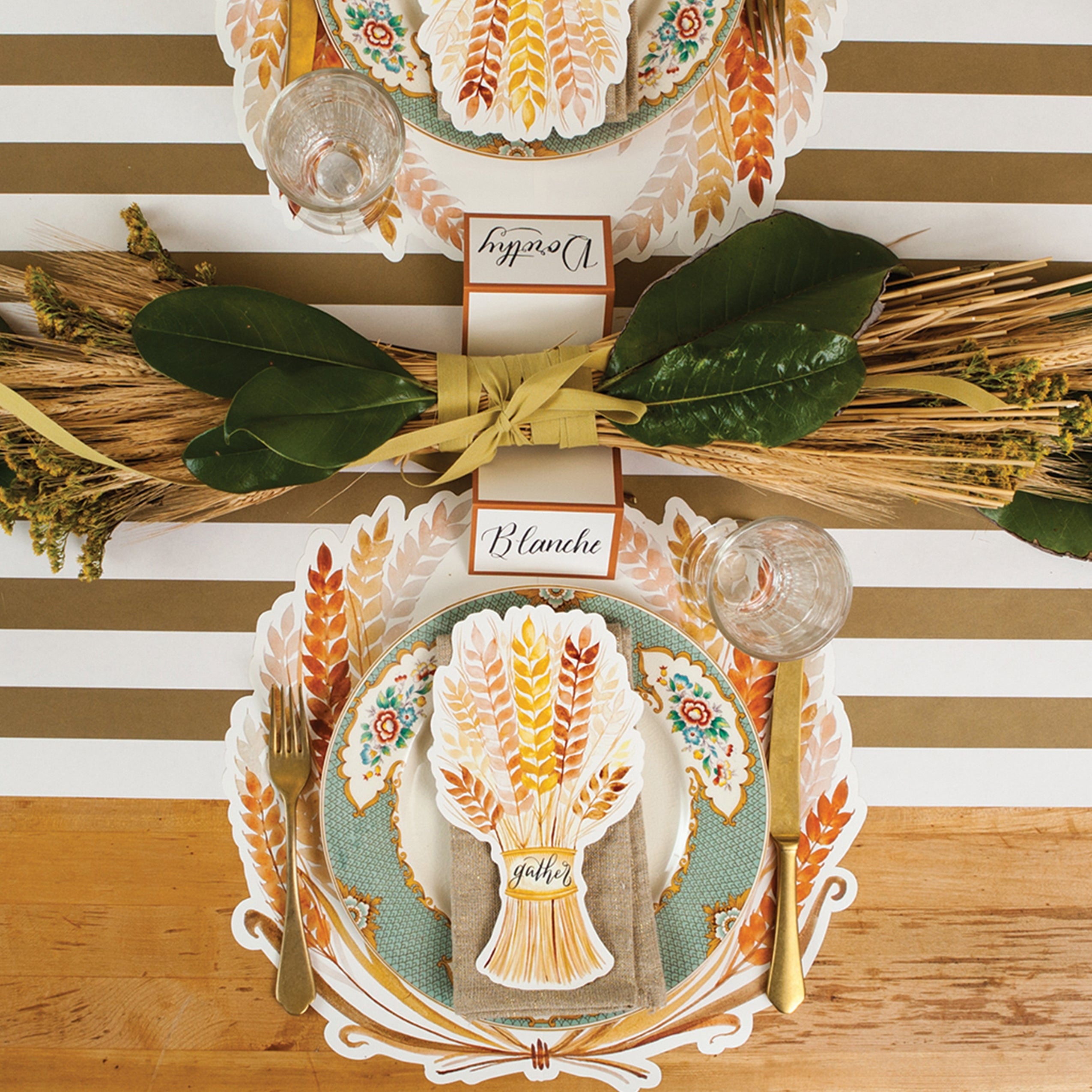 A top-down view of an elegant table setting for two featuring a Golden Harvest Table Accent labeled with a name resting on each plate.