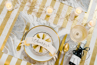An elegant gold and white place setting, featuring a Classic Gold Banner Table Accent with &quot;Cheers&quot; written on it  resting on the plate.