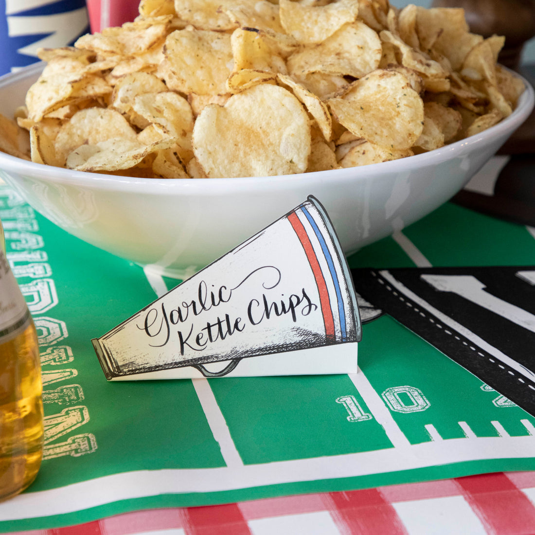 A Megaphone Place Card labeled &quot;Garlic Kettle Chips&quot; on a gameday snack table.