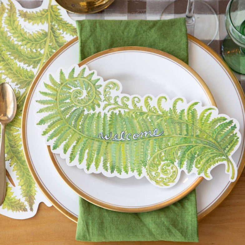 A Fern Fronds Table Accent with "Welcome" written on it resting on the plate of an elegant place setting, from above.