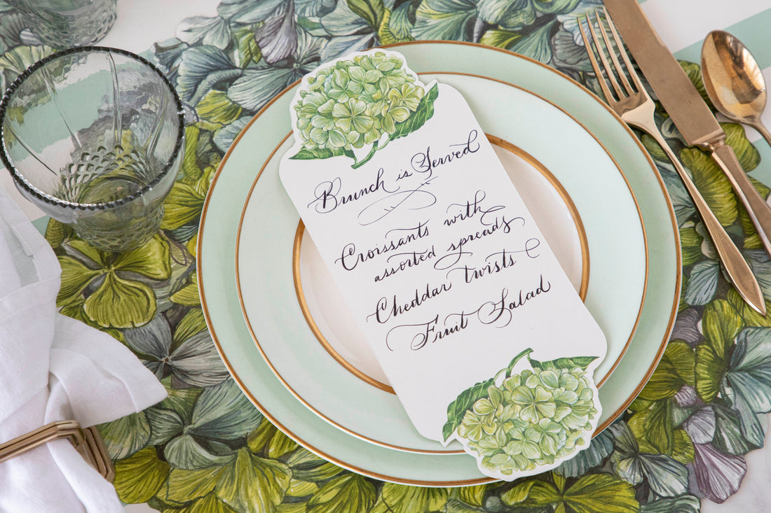 A Hydrangea Table Card with a brunch menu written on it resting on the plate of an elegant floral place setting.