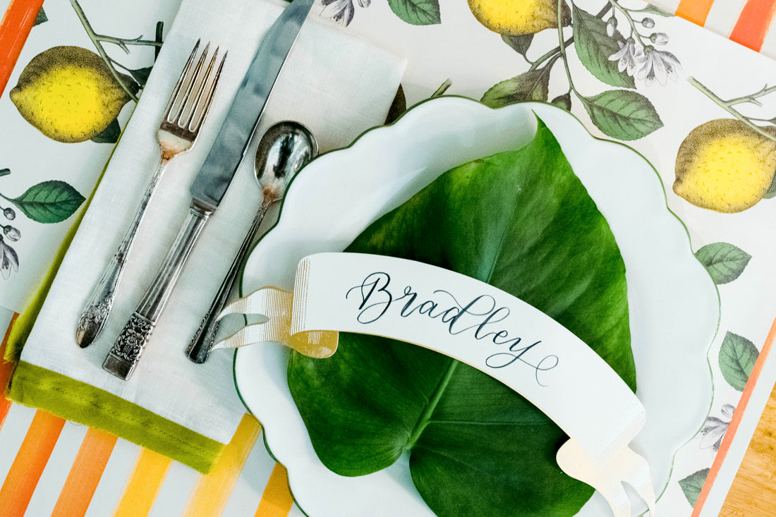 A top-down view of an elegant place setting featuring a Classic Gold Banner Table Accent labeled &quot;Bradley&quot; resting on the plate.