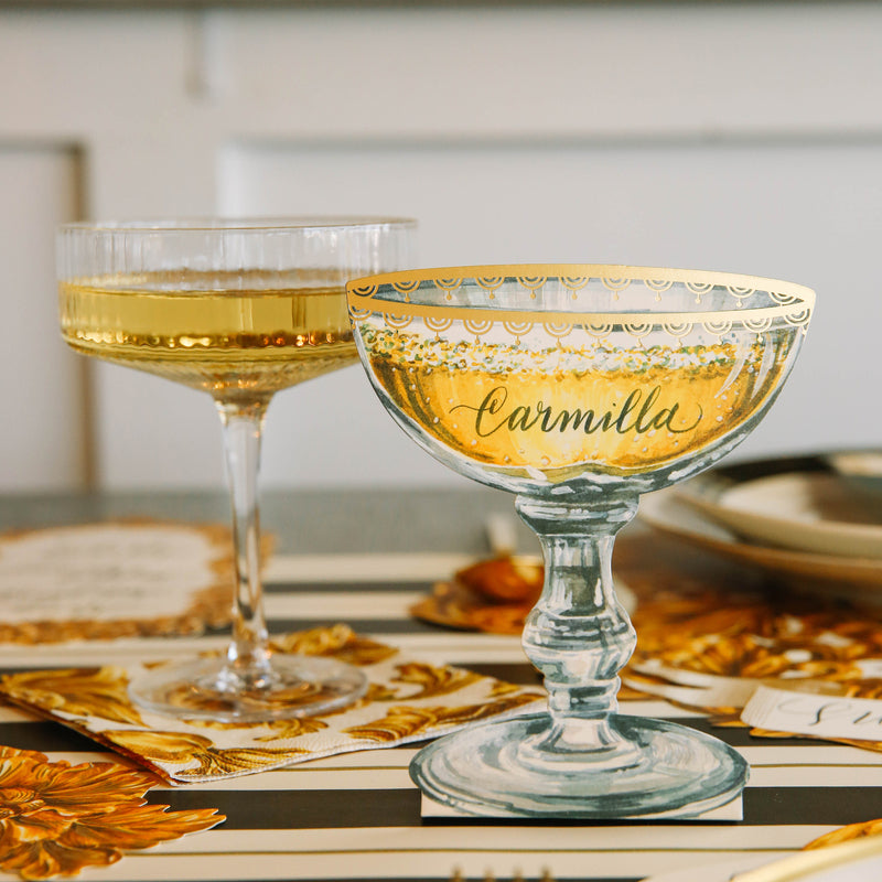 An elegant table setting with a Champagne Coupe Place Card reading "Carmilla" standing next to a real coupe of champagne.