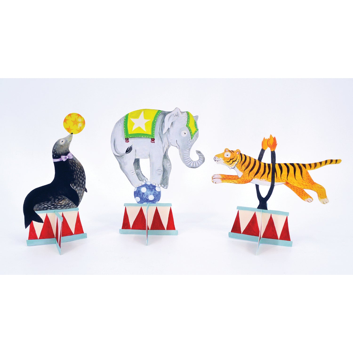 Three illustrated, die-cut paper ornaments that stand up on the table: a seal with a ball on its nose, an elephant balancing on a ball, and a tiger jumping through a flaming hoop.