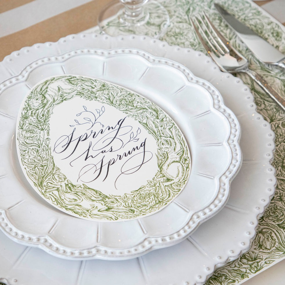 A Greenhouse Hares Table Card with "Spring has Sprung" written on it in elegant script resting on the plate of a Springtime place setting.