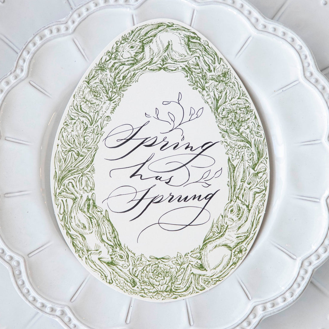 Close-up of a Greenhouse Hares Table Card with &quot;Spring has Sprung&quot; written on it in elegant script resting on a white plate.