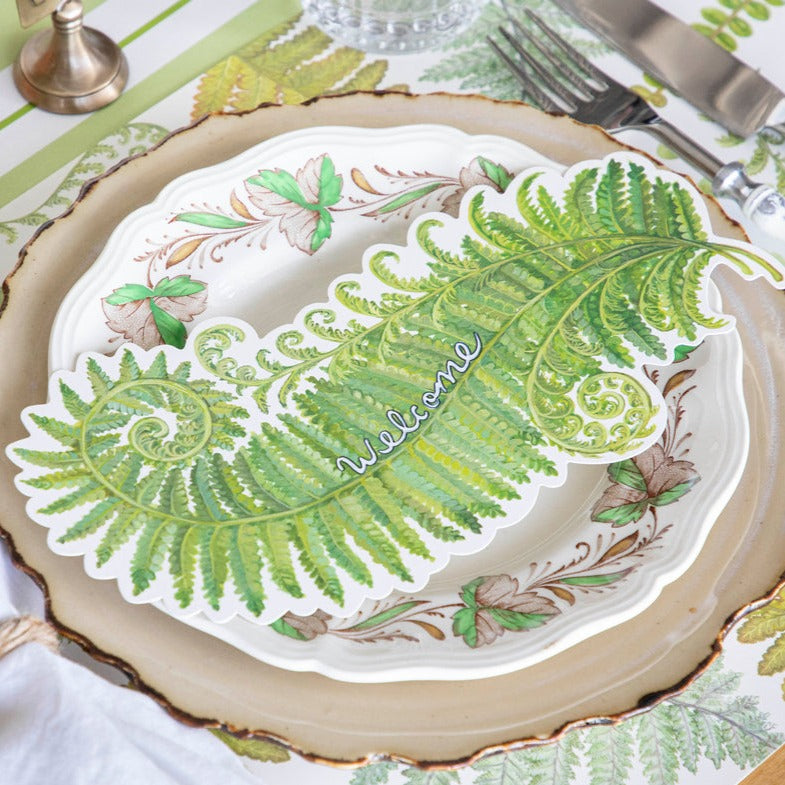 A Fern Fronds Table Accent with "Welcome" written on it resting on the plate of an elegant place setting.