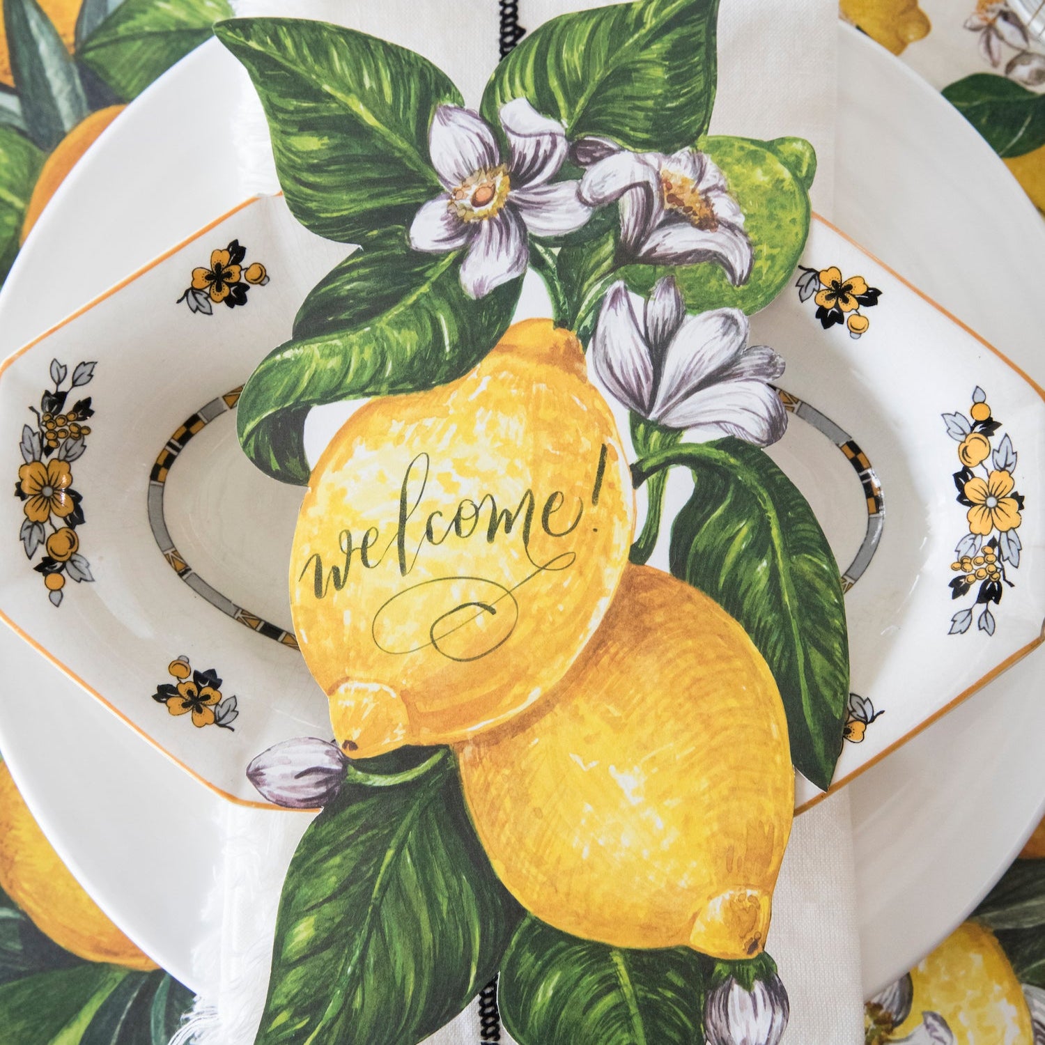 A vibrant citrus-themed place setting featuring a Lemon Table Accent with "Welcome!" written on it resting on the plate.