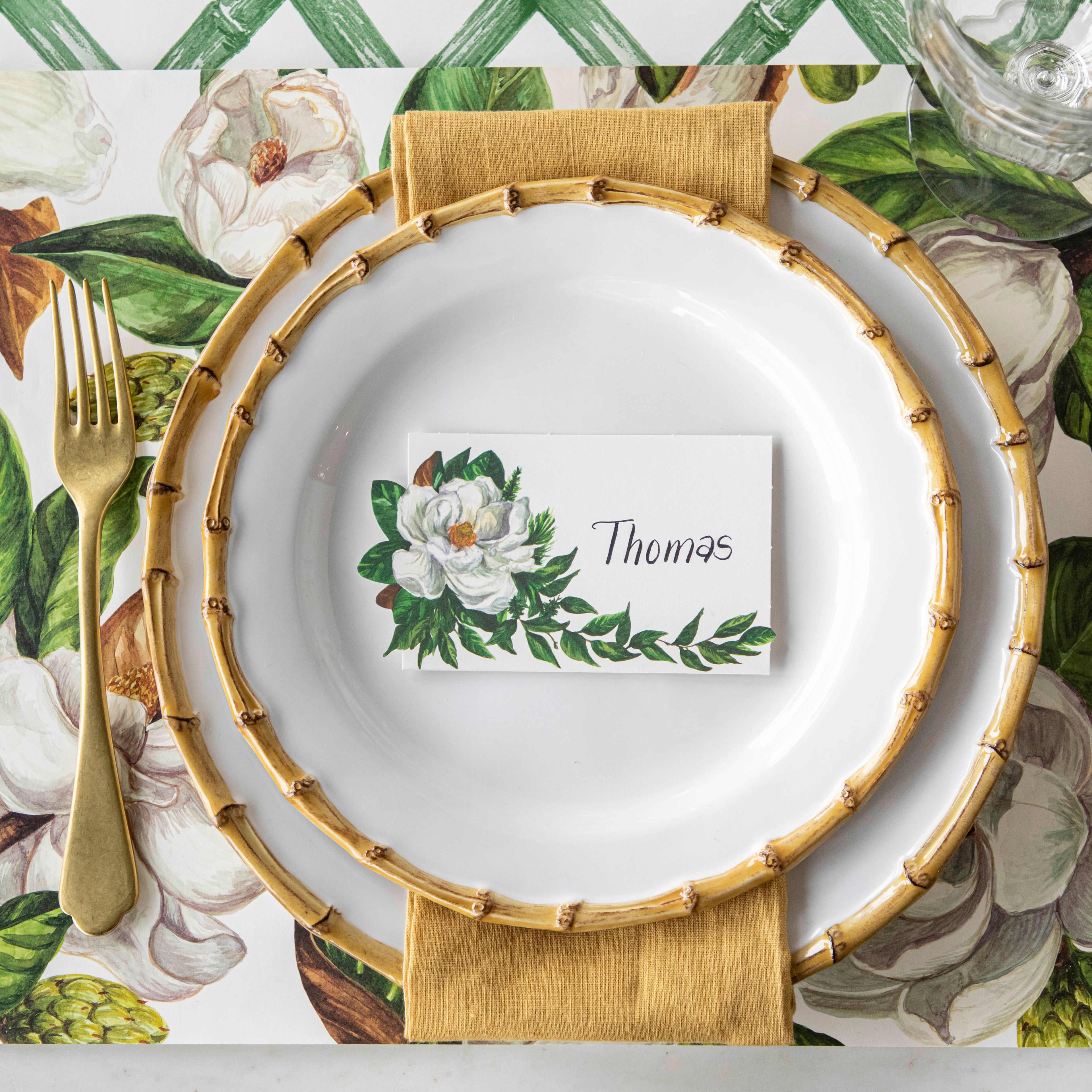 Top-down view of an elegant place setting featuring a Magnolia Place Card labeled "Thomas" laying flat on the plate.
