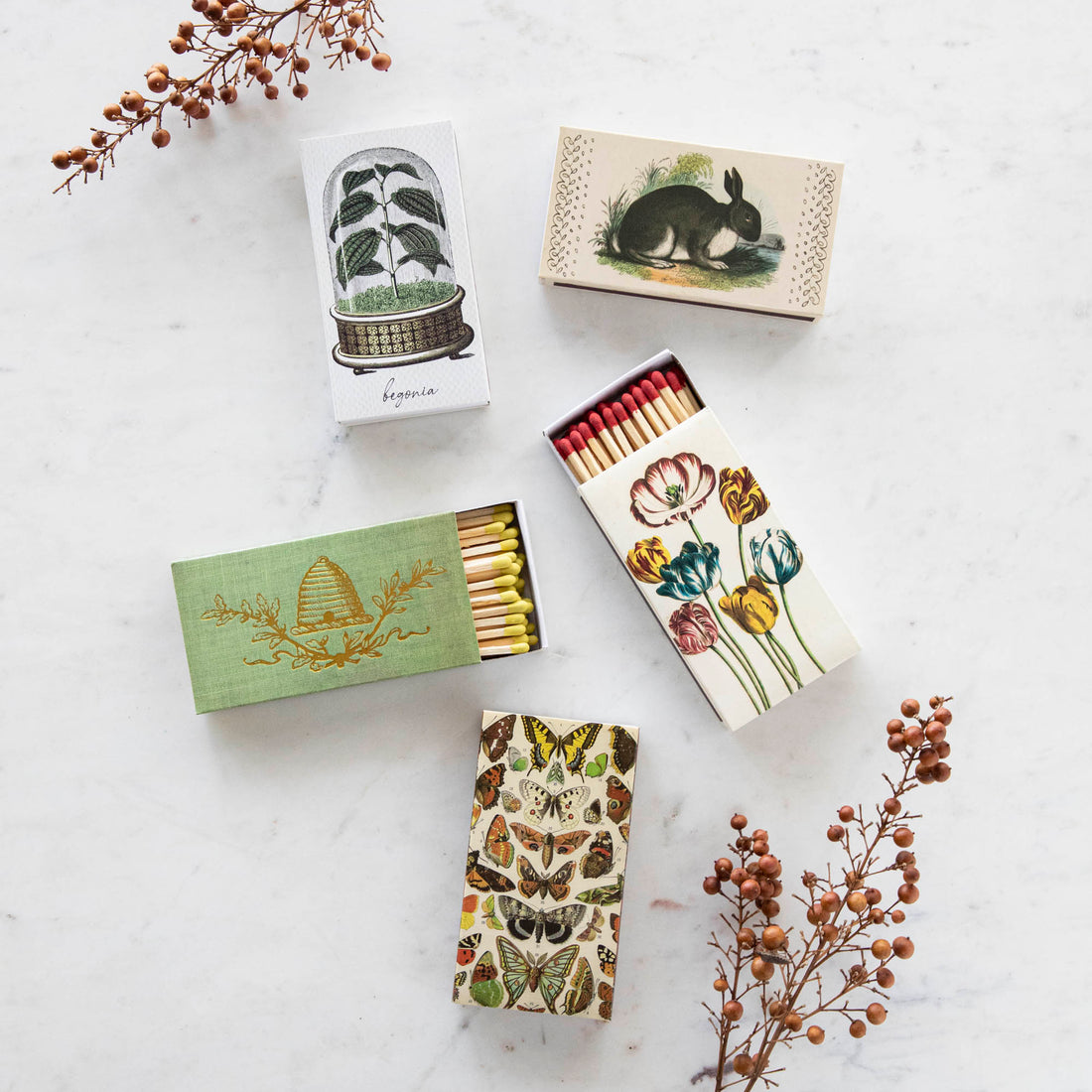 Four boxes of Nature Matches by HomArt with decorative floral and botanical designs laid out on a marble surface, accompanied by sprigs of dried plants.