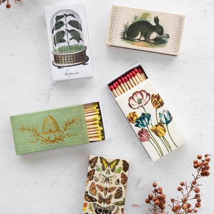 Four boxes of Nature Matches by HomArt with decorative floral and botanical designs laid out on a marble surface, accompanied by sprigs of dried plants.