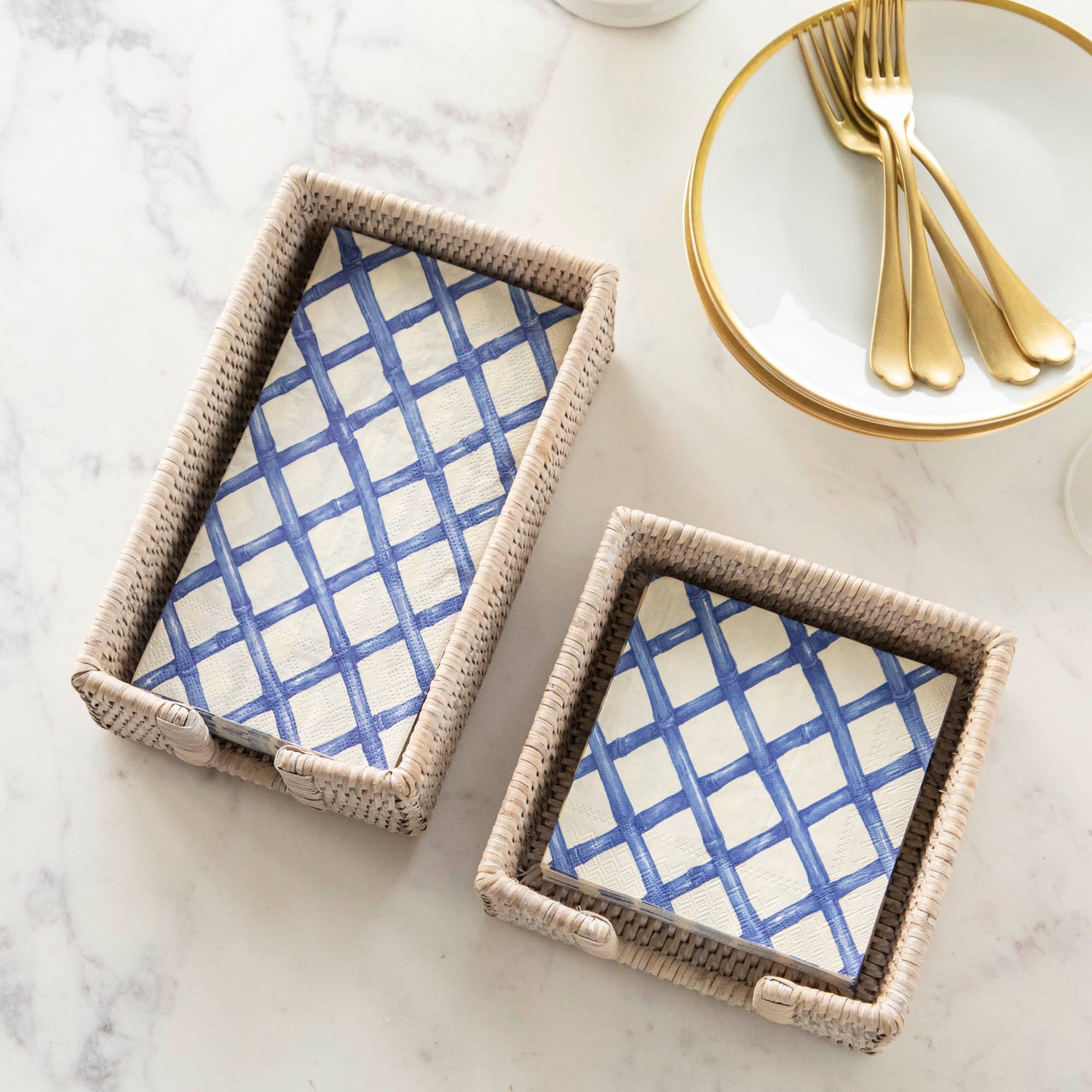 A stack of Blue Lattice Guest Napkins and a stack of Blue Lattice Cocktail Napkins in wicker napkin holders, next to a stack of plates with gold forks.