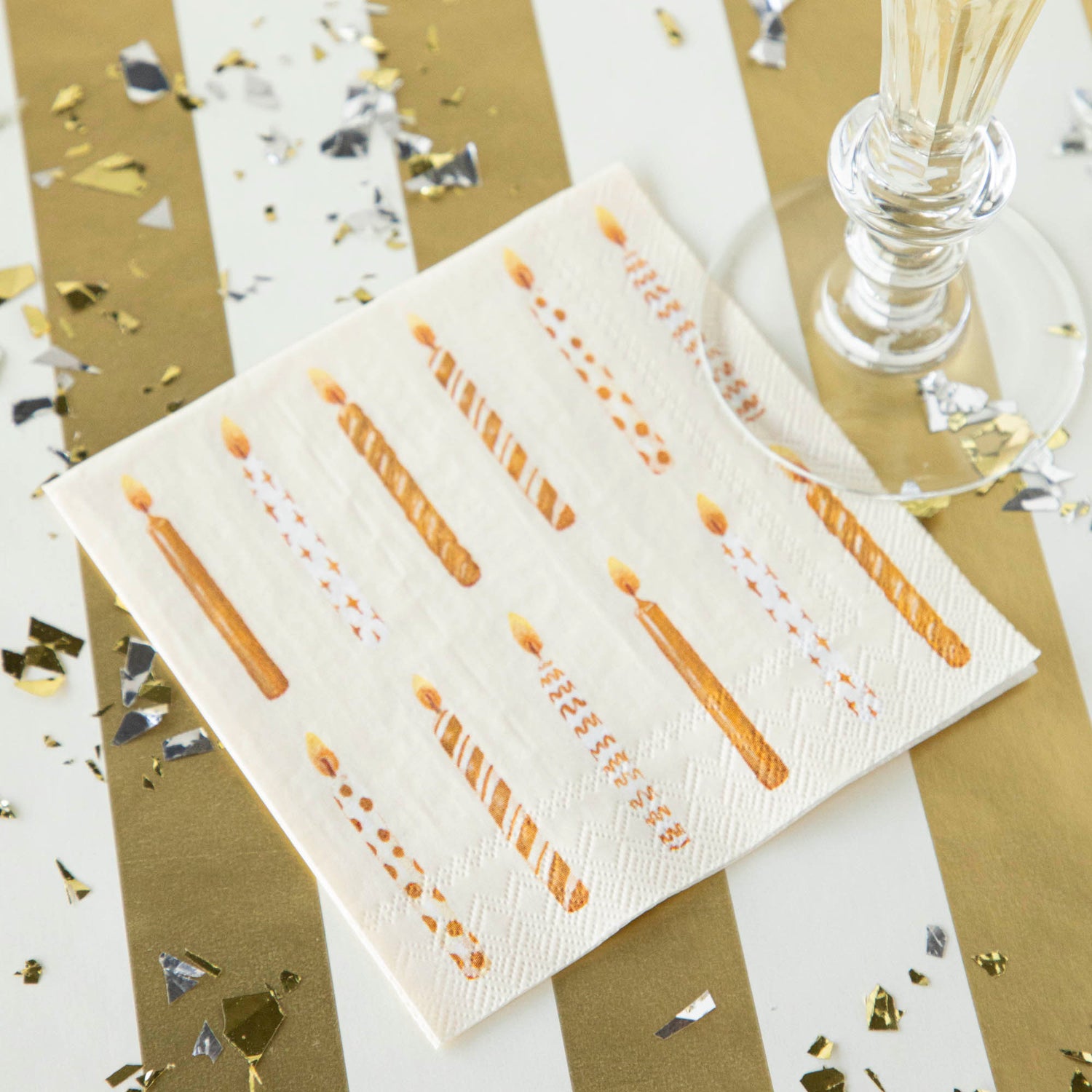 A Gold Candles Cocktail Napkin under a stemmed glass on a festive gold and white table.