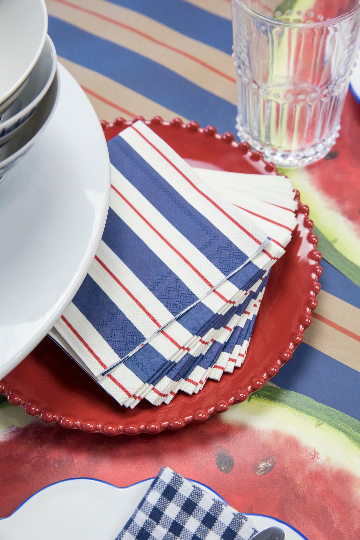 A stack of Navy & Red Awning Stripe Cocktail Napkins fanned out on a red plate in a summertime table setting.