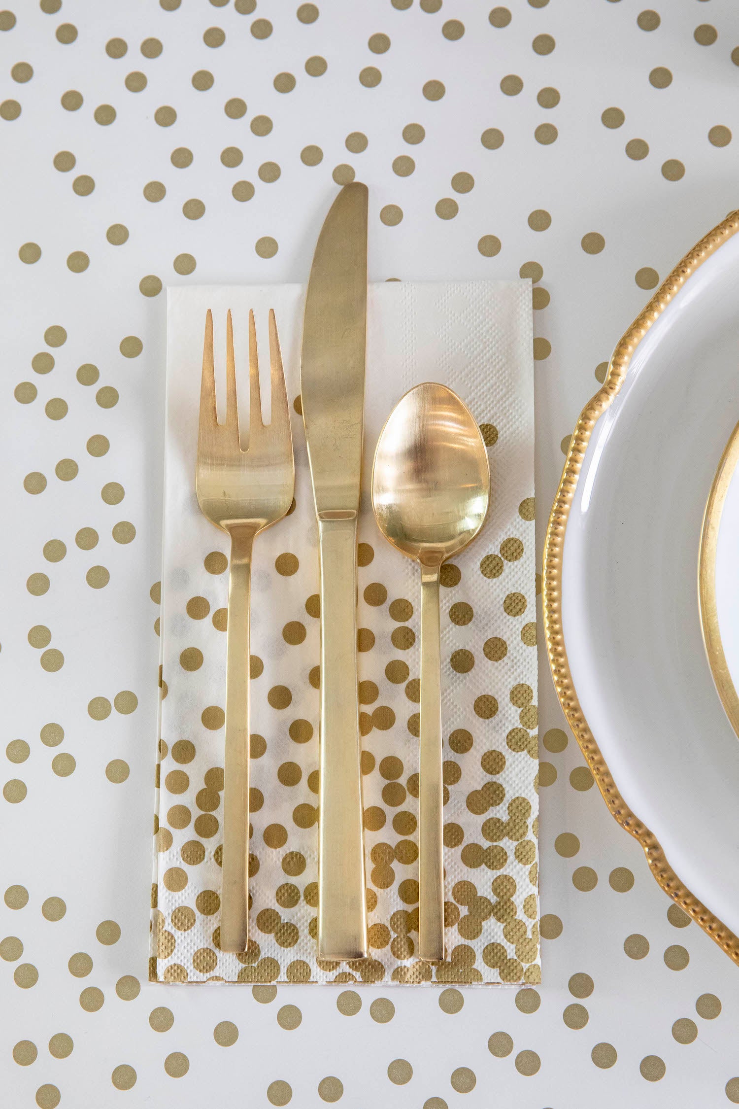 A Gold Confetti Guest napkin under gold cutlery next to a gold-rimmed plate, on a gold confetti table runner.